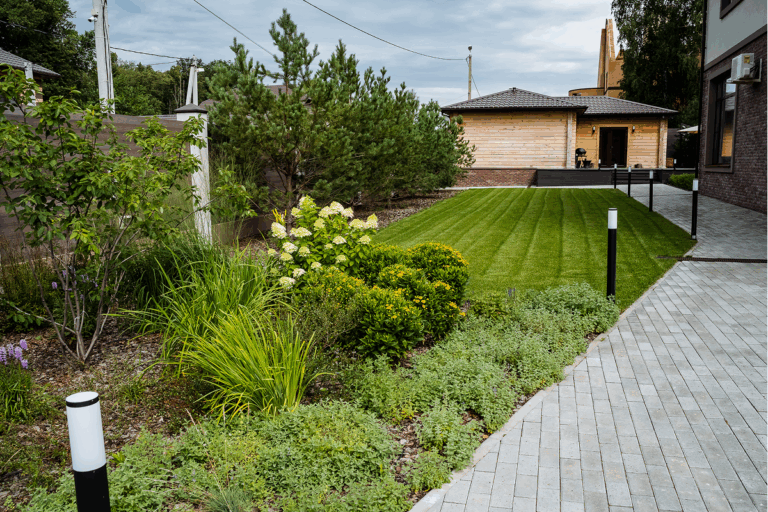 Modern landscaped backyard with lush lawn, flower beds, pine trees, stone walkway, and wooden guesthouse.